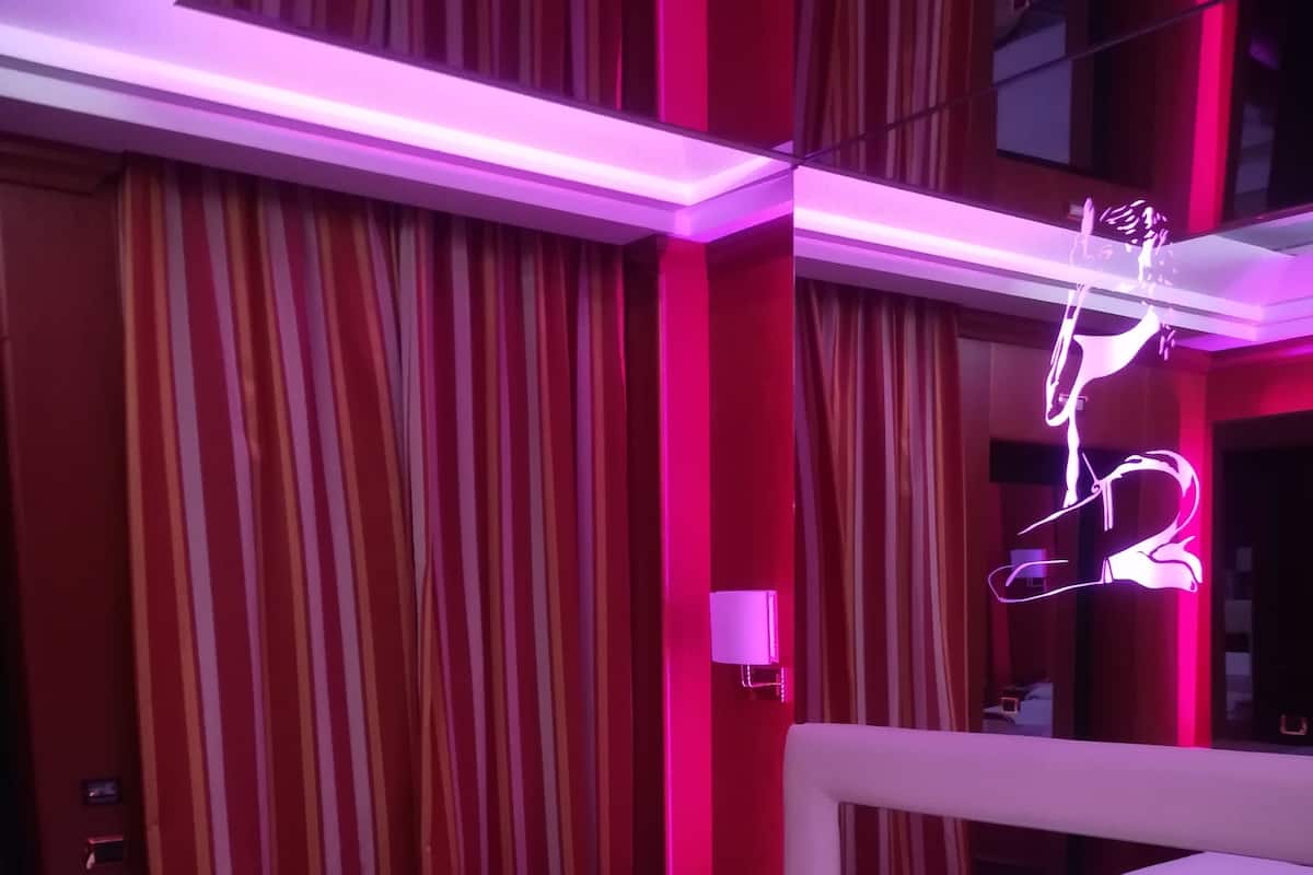 Una habitación de hotel con una cama, cortinas a rayas, un letrero de neón y una mesita de noche.