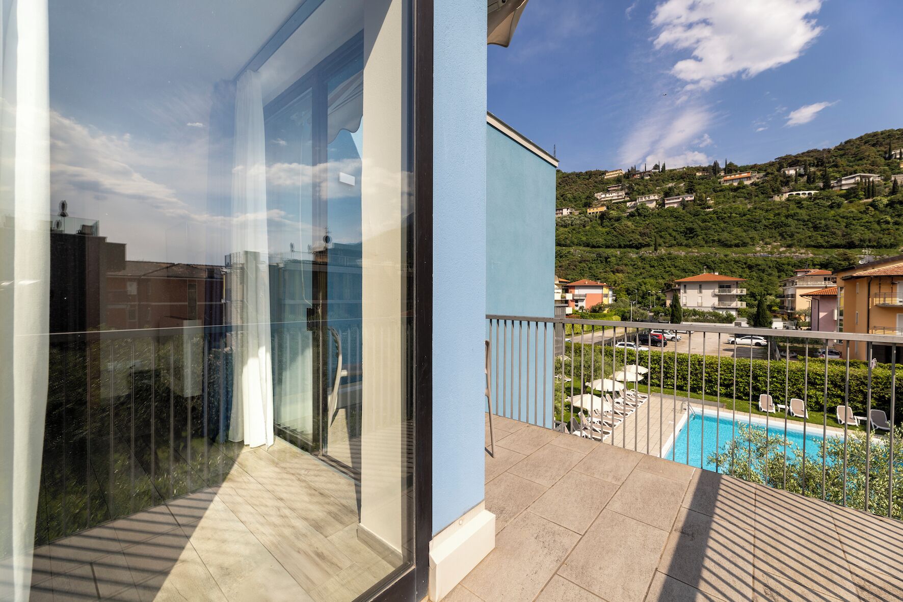 A balcony with a glass door, a pool, and a view of a hillside with houses.