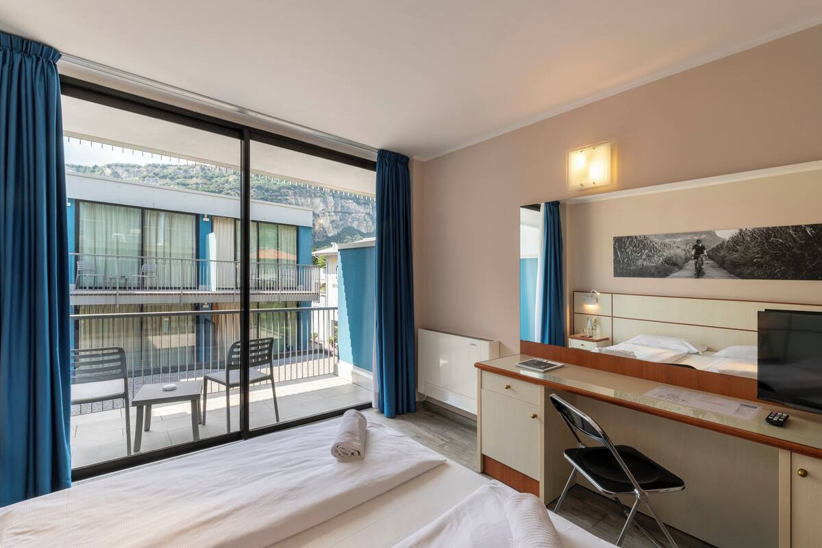 A hotel room with a bed, a desk, a chair, a TV, and a balcony with a view of the mountains.