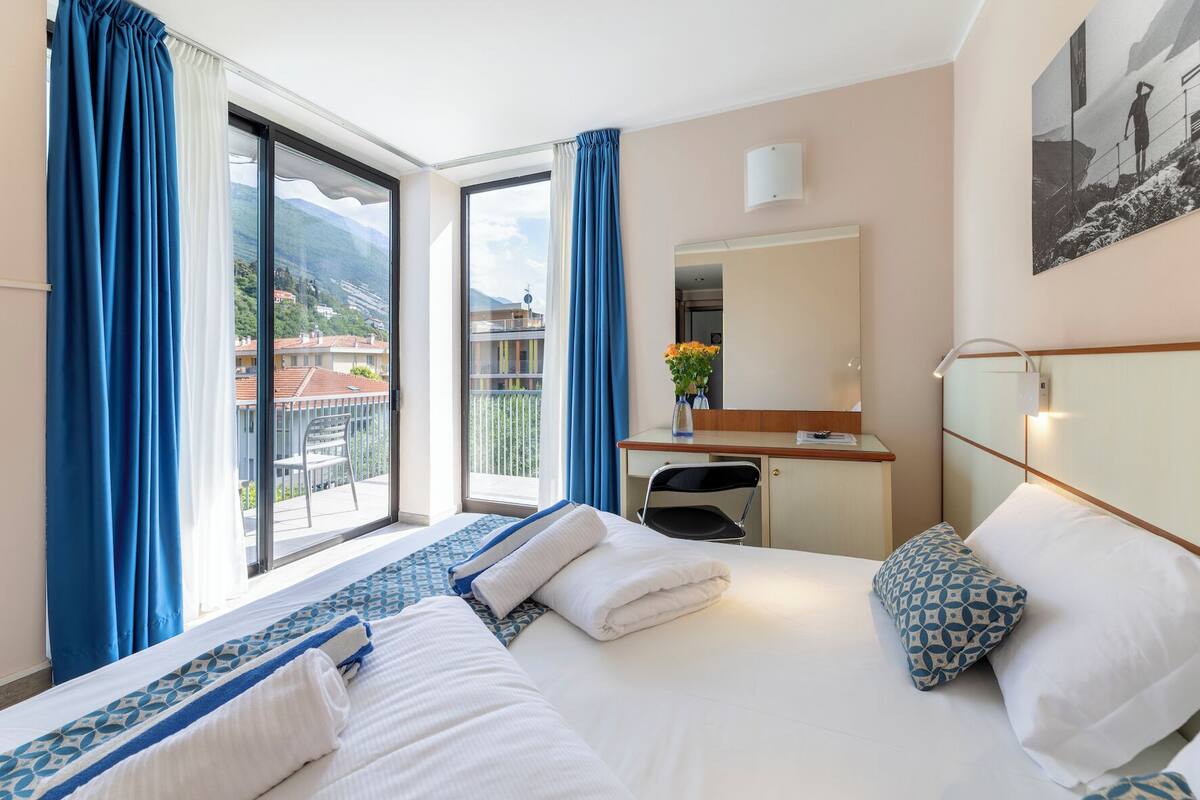 A hotel room with a large bed, a desk, a chair, and a balcony with curtains.