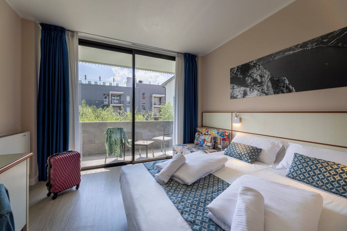 A hotel room with a large bed, a small table, a chair, a suitcase, and a balcony with a view of buildings and greenery.