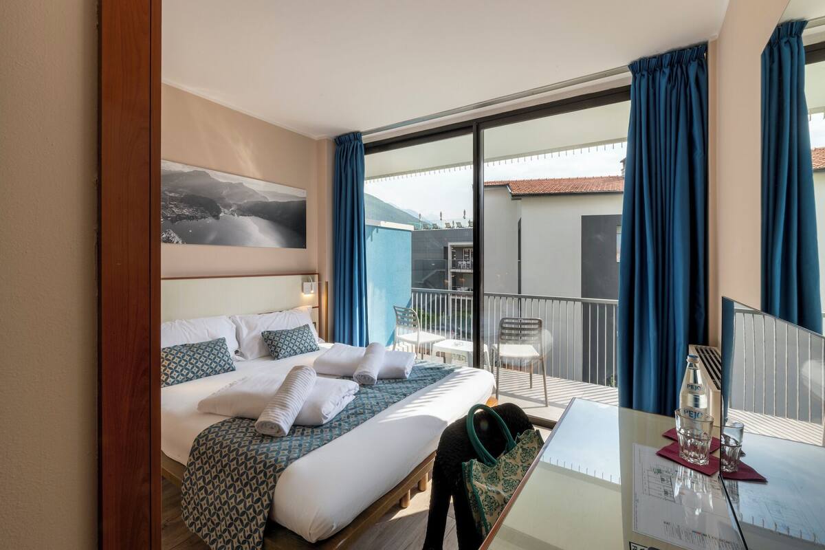 A hotel room with a bed, a desk with a computer, a chair, and a balcony with a view of buildings and mountains.