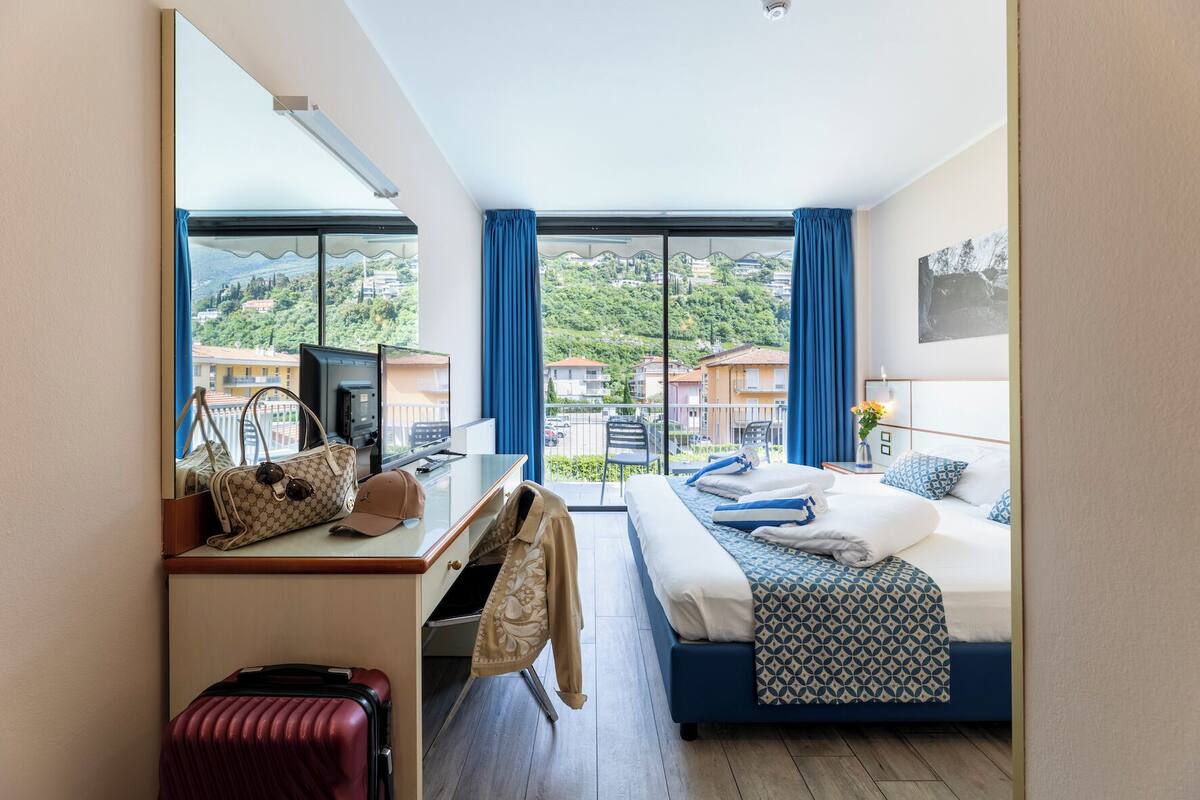 A hotel room with a bed, a desk, a chair, a suitcase, and a view of the city through large windows.
