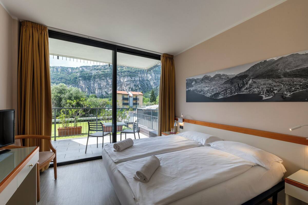 A hotel room with two beds, a TV, a desk, and a balcony with a view of greenery and a bridge.