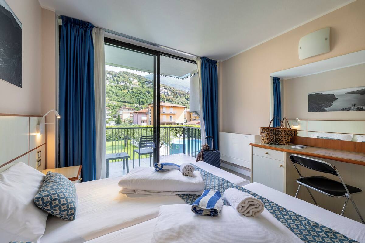 A hotel room with two beds, a desk, a chair, a view of a balcony with a pool, and a cityscape in the background.