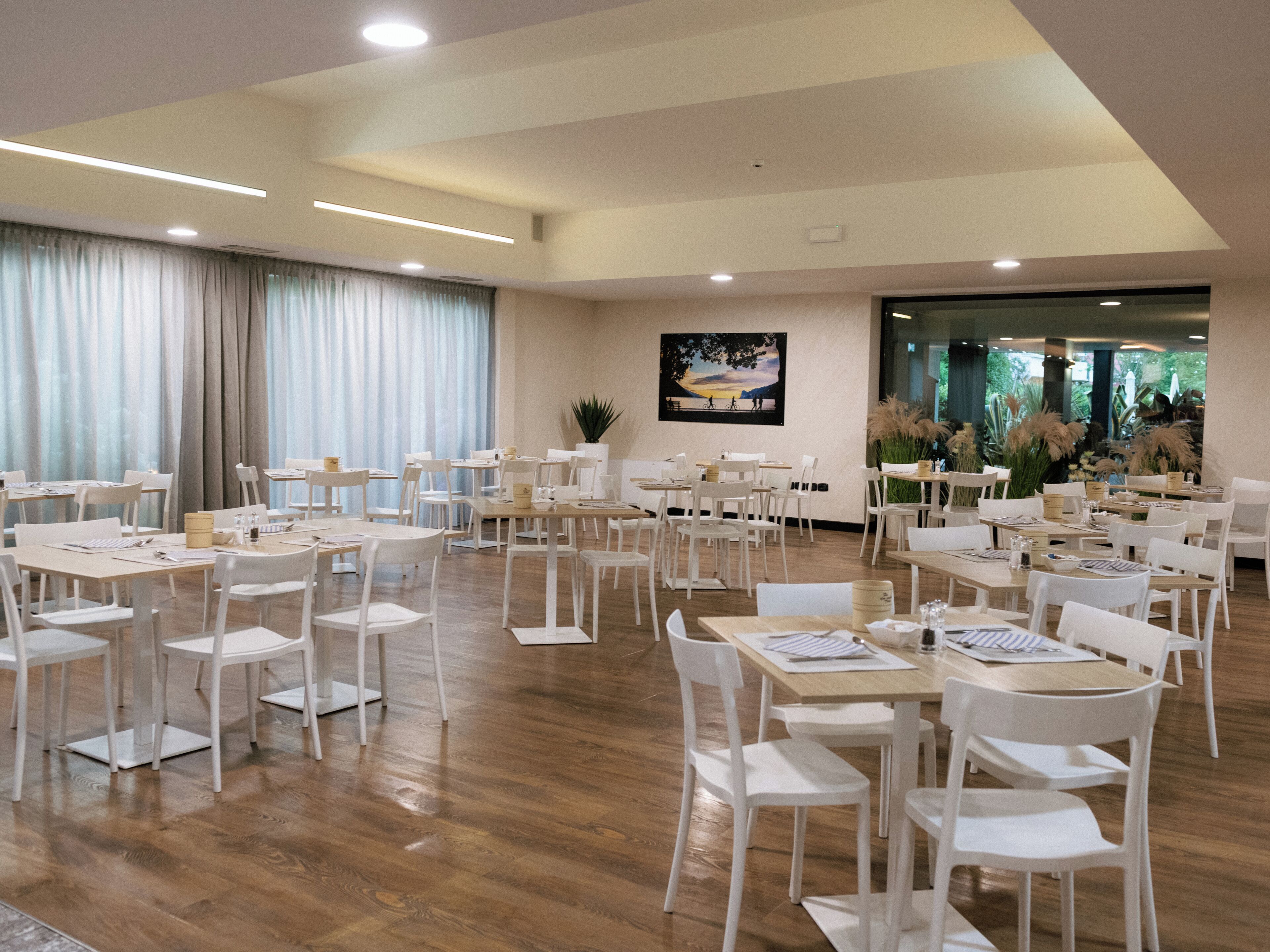 A spacious dining area with white tables and chairs, a large window with curtains, and a painting on the wall.