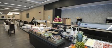 Zona de bufé con diversas opciones de alimentos, como frutas, carnes y platos ya preparados, en un entorno moderno de hotel.