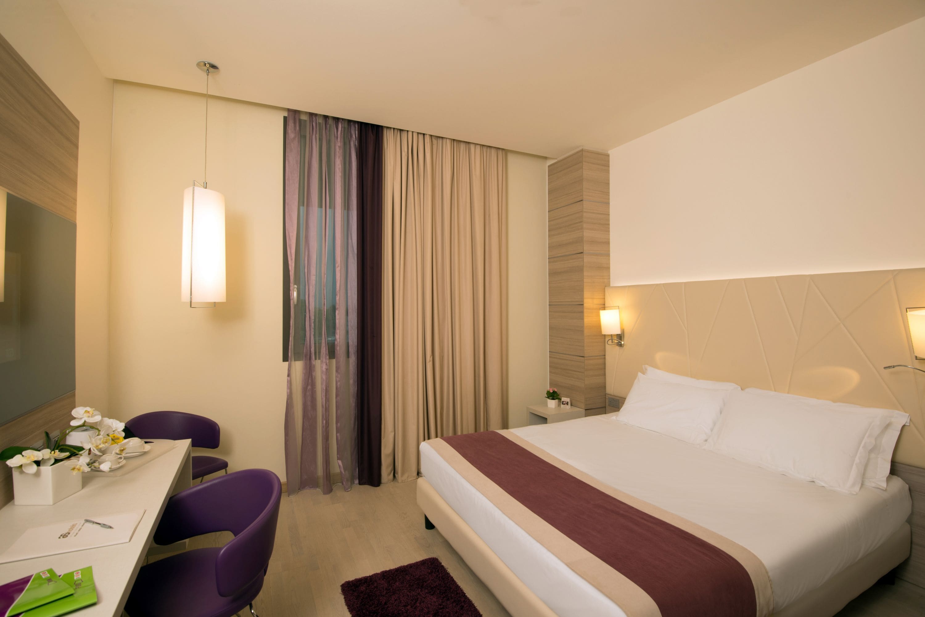 A hotel room with a bed, a desk with chairs, a lamp, and a curtain.