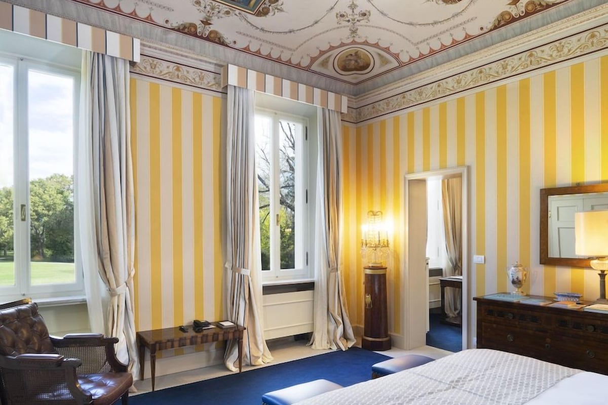 A room with a large bed, a chair, a desk, a lamp, and a mirror. There are curtains on the windows, and the walls have a striped pattern.