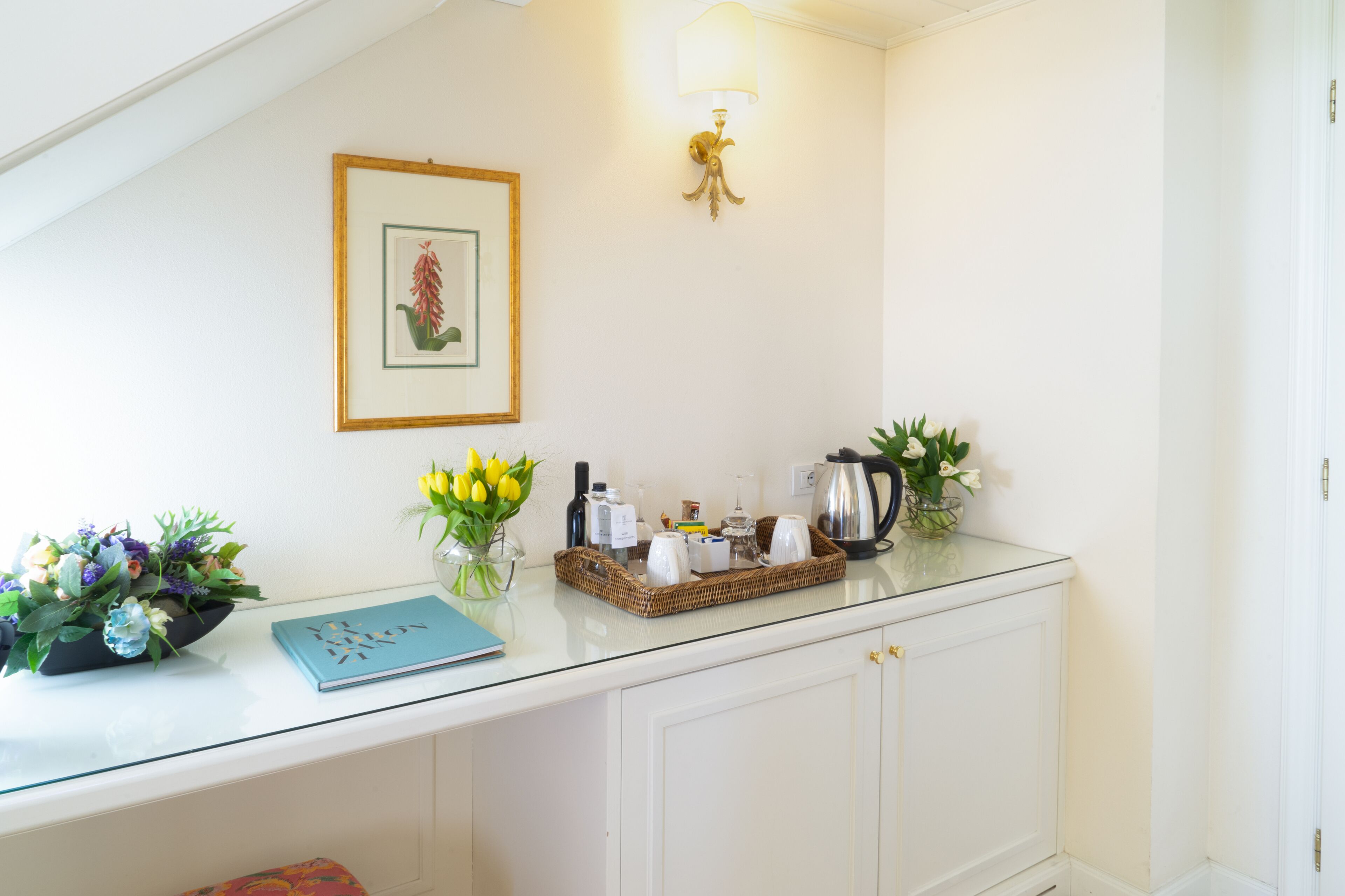 A room with a glass-top table, a framed botanical illustration, a vase with yellow flowers, a wicker tray, and a white cabinet.