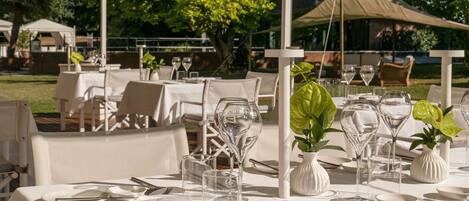 An outdoor dining area with tables set for a meal, surrounded by greenery and a tent structure.