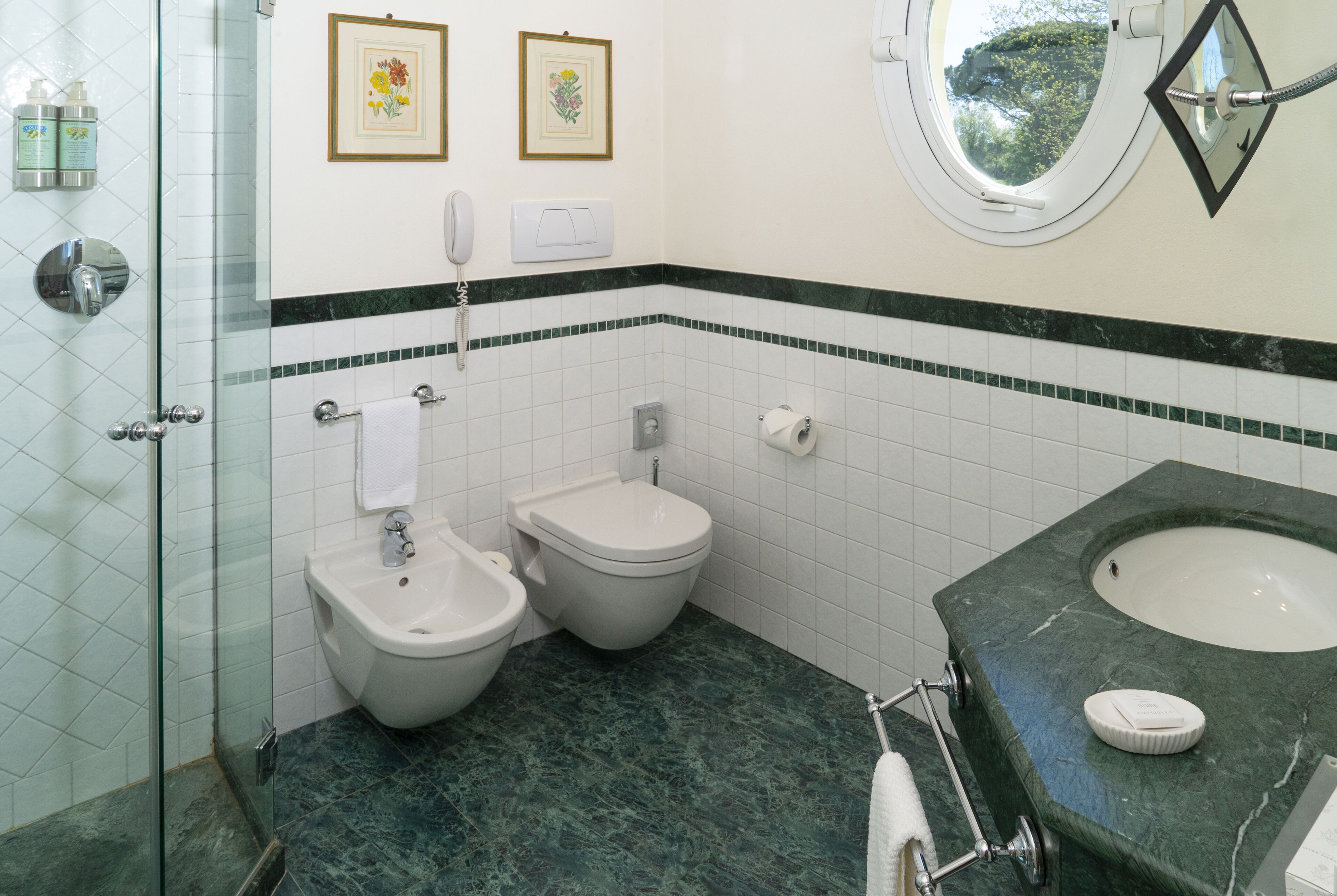 Uma casa de banho com cabine de duche em vidro, dois bidés, uma pia com bancada de mármore e uma janela com vista para árvores.