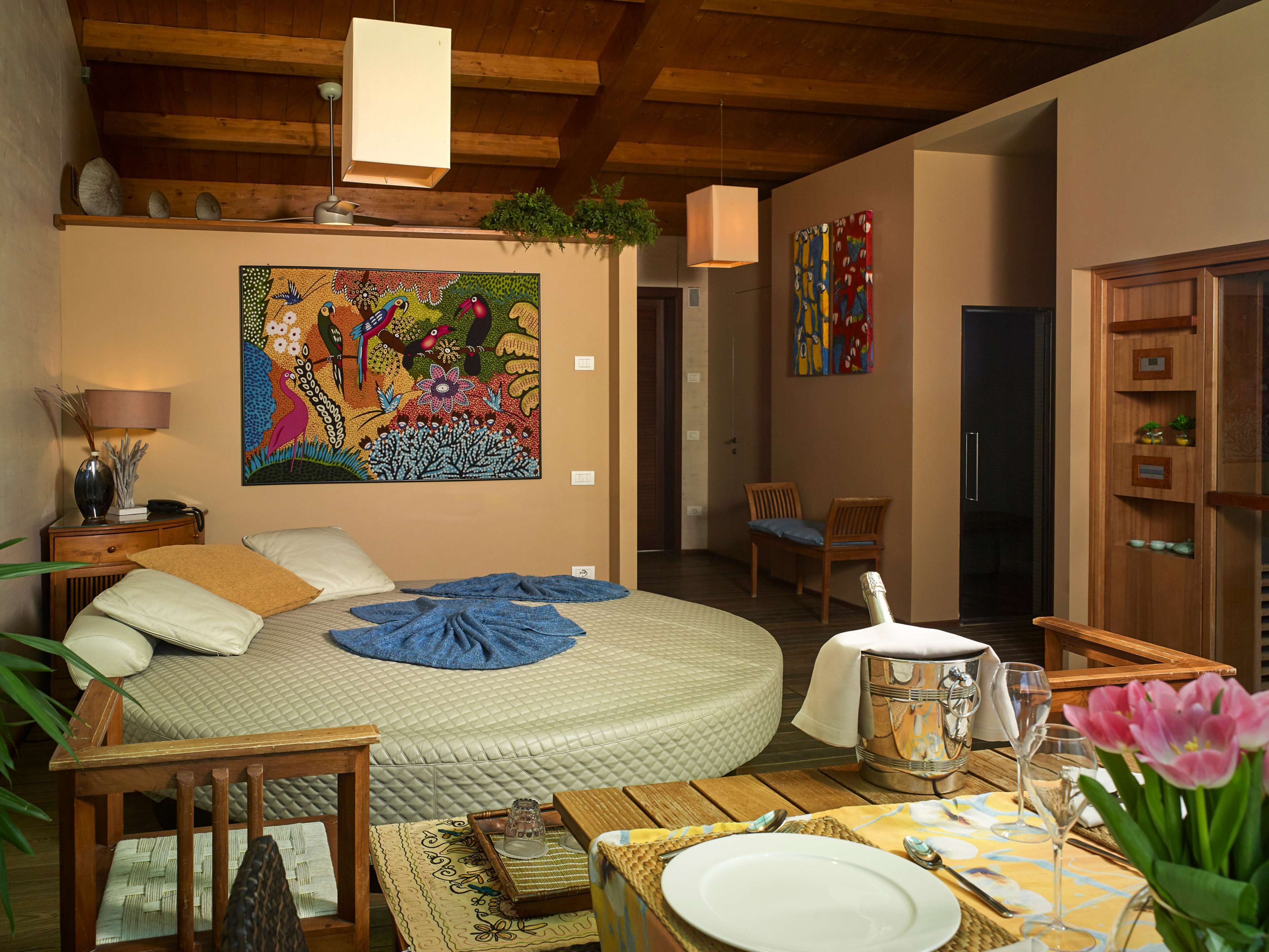 Une chambre confortable avec un lit rond, une table à manger dressée pour deux personnes et un tableau abstrait aux couleurs vives accroché au mur.