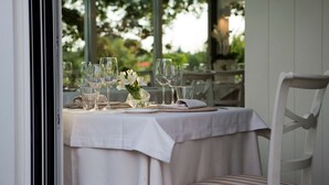 Una mesa lista para comer, con vasos transparentes y un jarrón de flores blancas, sobre un fondo de follaje.