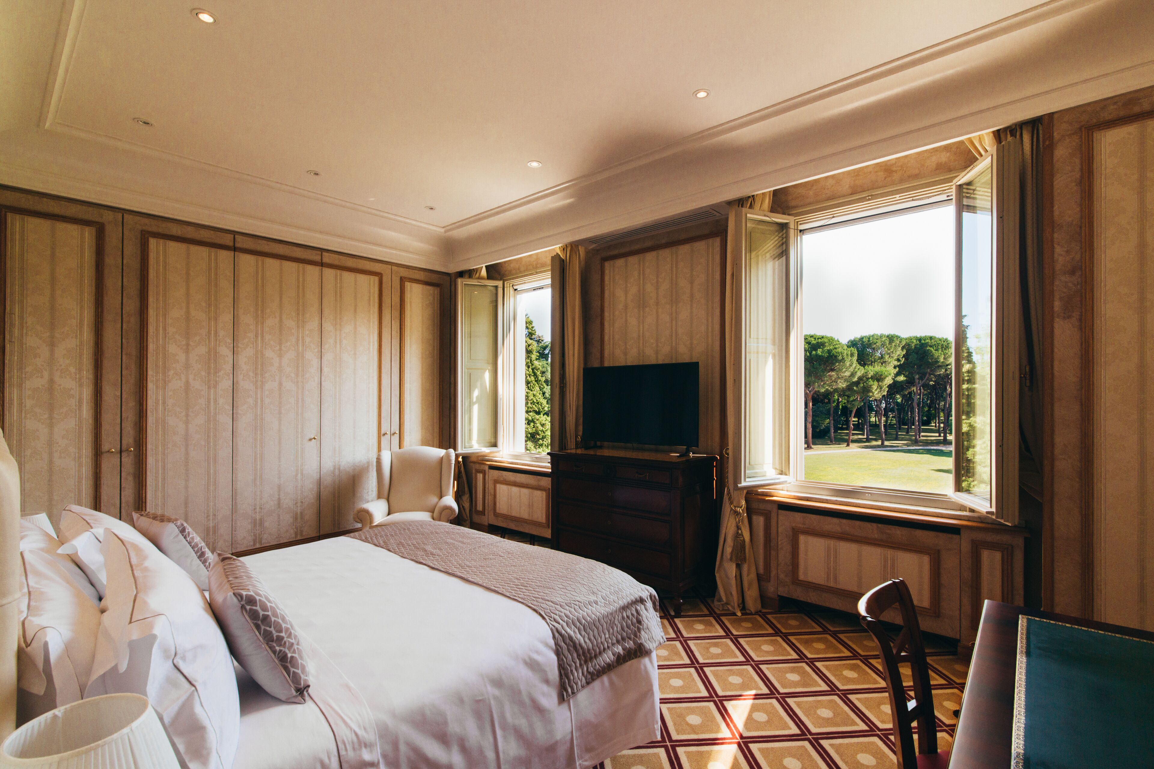 A hotel room with a large bed, a television, a chair, and a window with a view of trees.