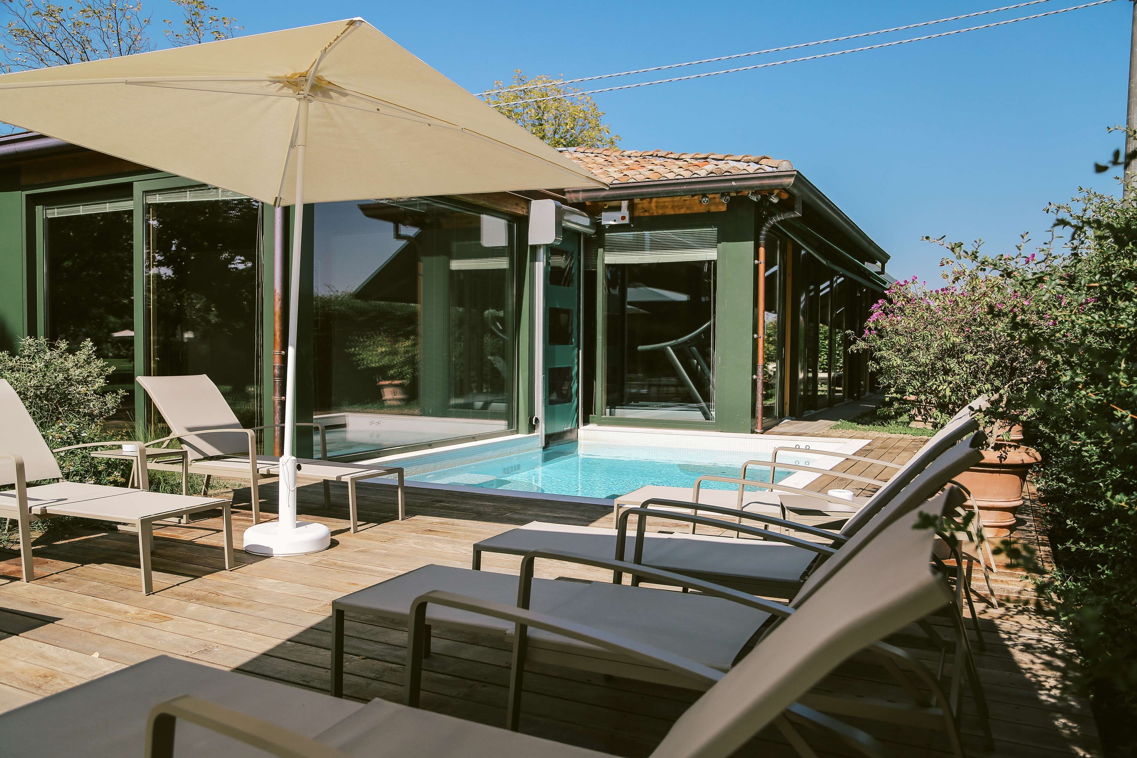 Zona de piscina amb gandules i un parasol gran, envoltada d'una terrassa de fusta i portes de vidre que donen a un edifici.