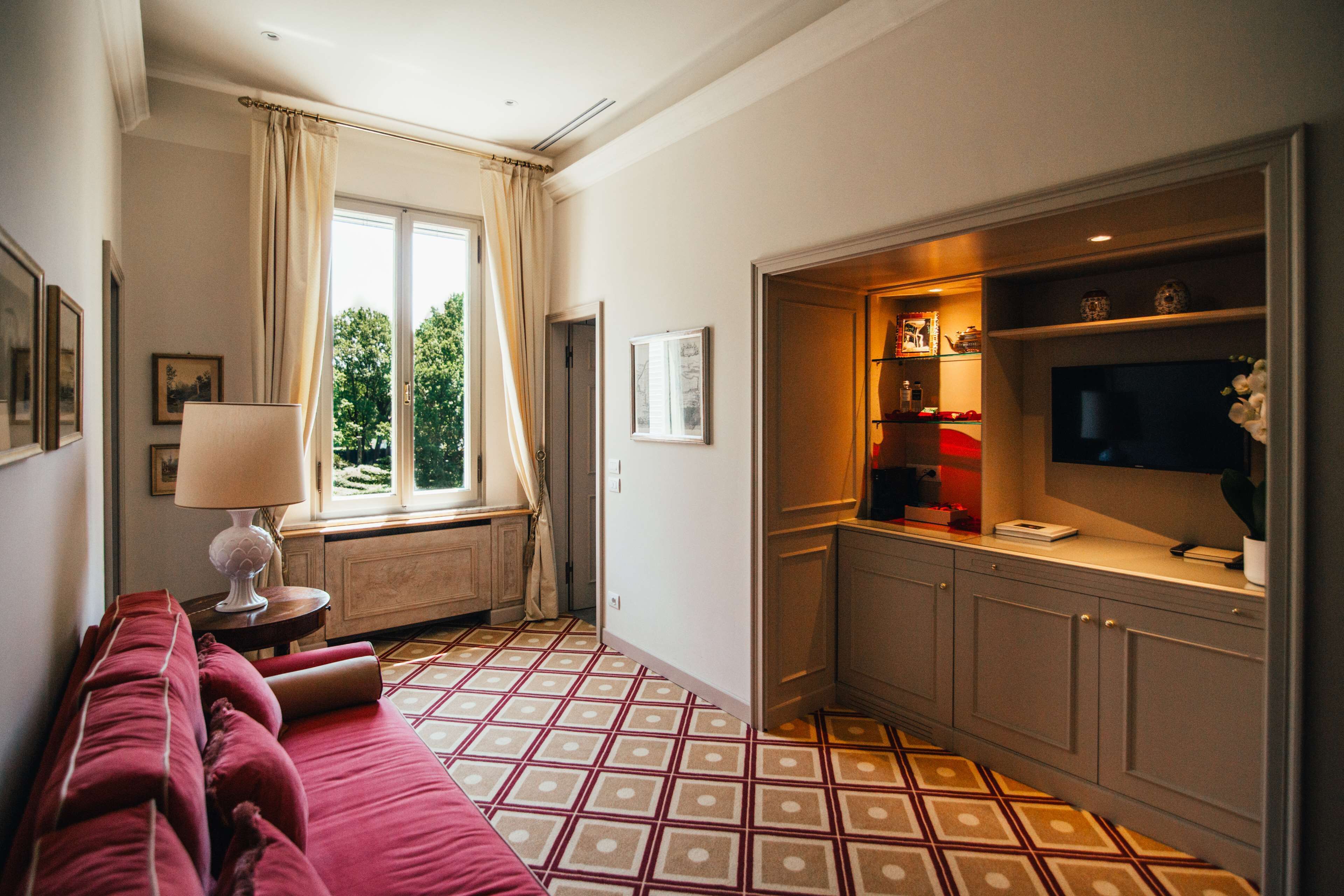Una habitación con sofá rojo, ventana con cortinas, zona de cocina con armarios y televisor, y suelo estampado.