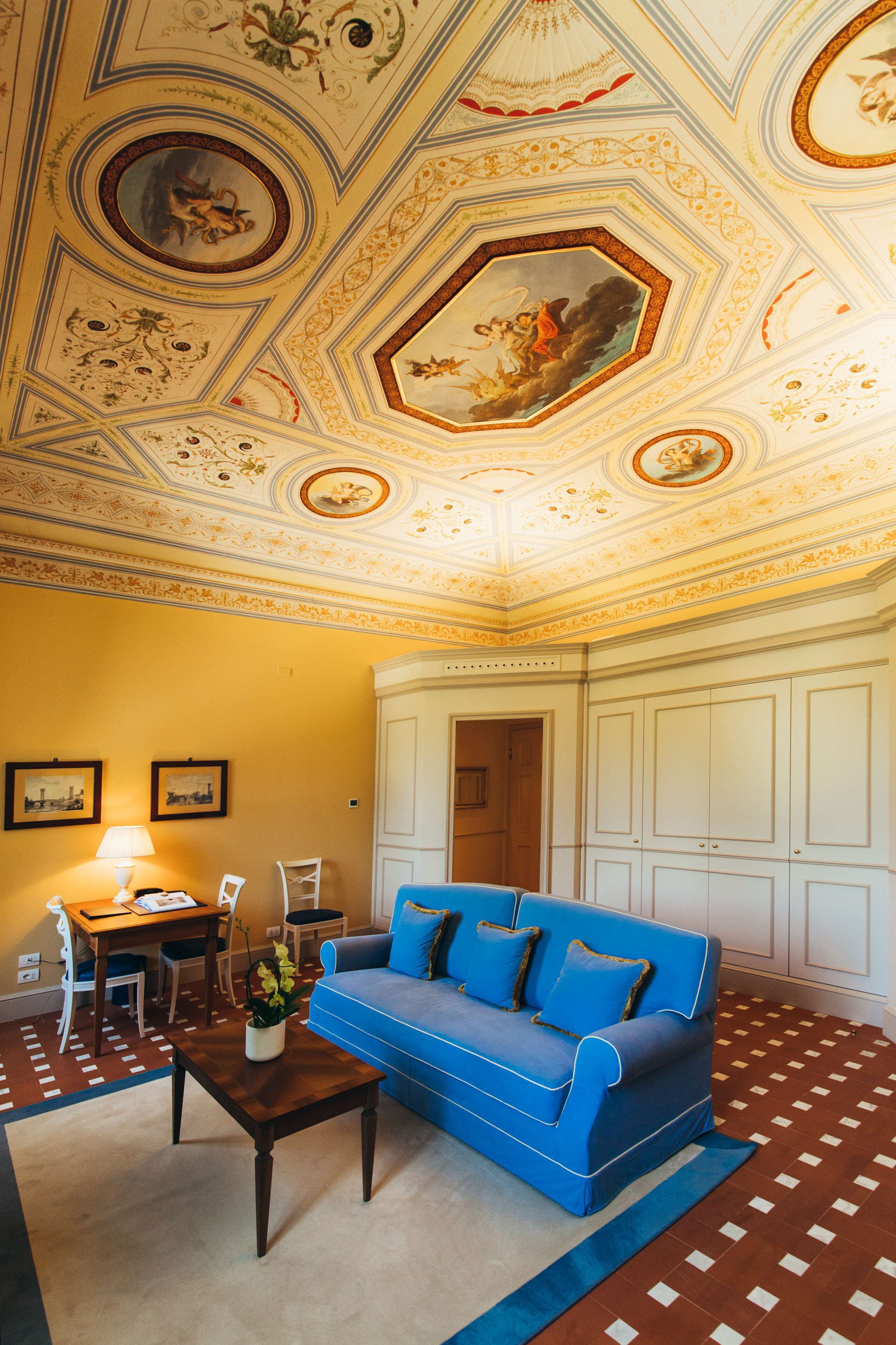 Ein Zimmer mit einem blauen Sofa, einem Holztisch und einem Schreibtisch mit Lampe. Die Decke zeigt aufwändige Verzierungen und Kunstwerke.