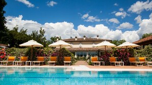 Una piscina con sillones de descanso y sombrillas, rodeada por un edificio con techo de tejas y un jardín lleno de flores.