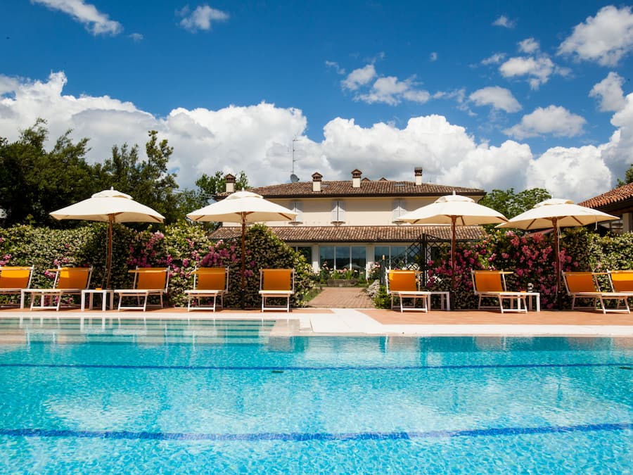 A swimming pool with lounge chairs and umbrellas, surrounded by a building with a tiled roof and a garden with flowers.