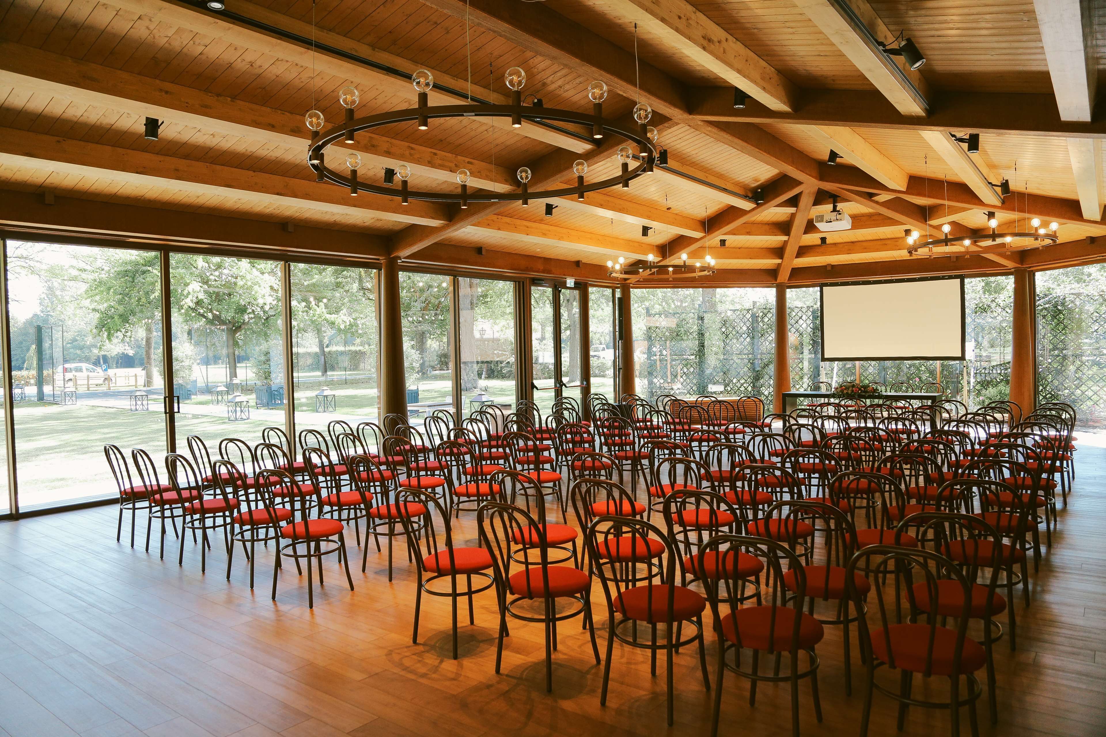 A large hall with rows of chairs and a projector screen, wooden ceiling beams, and large windows.