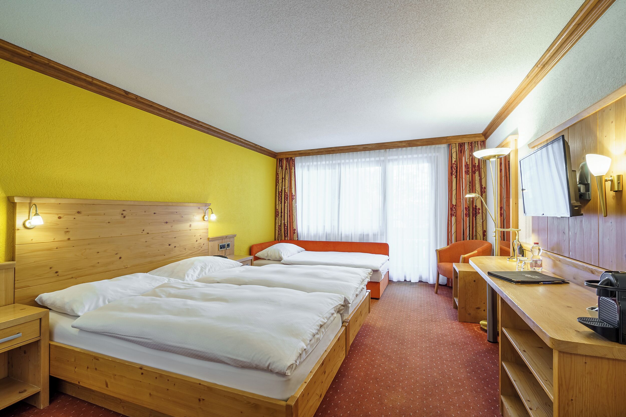 Ein Hotelzimmer mit zwei Holzbett, einem Schreibtisch mit Stuhl, einem Spiegel und einem Fenster mit Vorhängen.