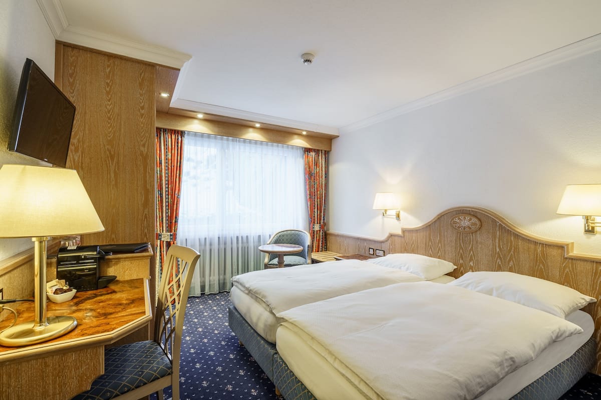 A hotel room with two beds, a desk with a lamp, a chair, a television, and a window with curtains.