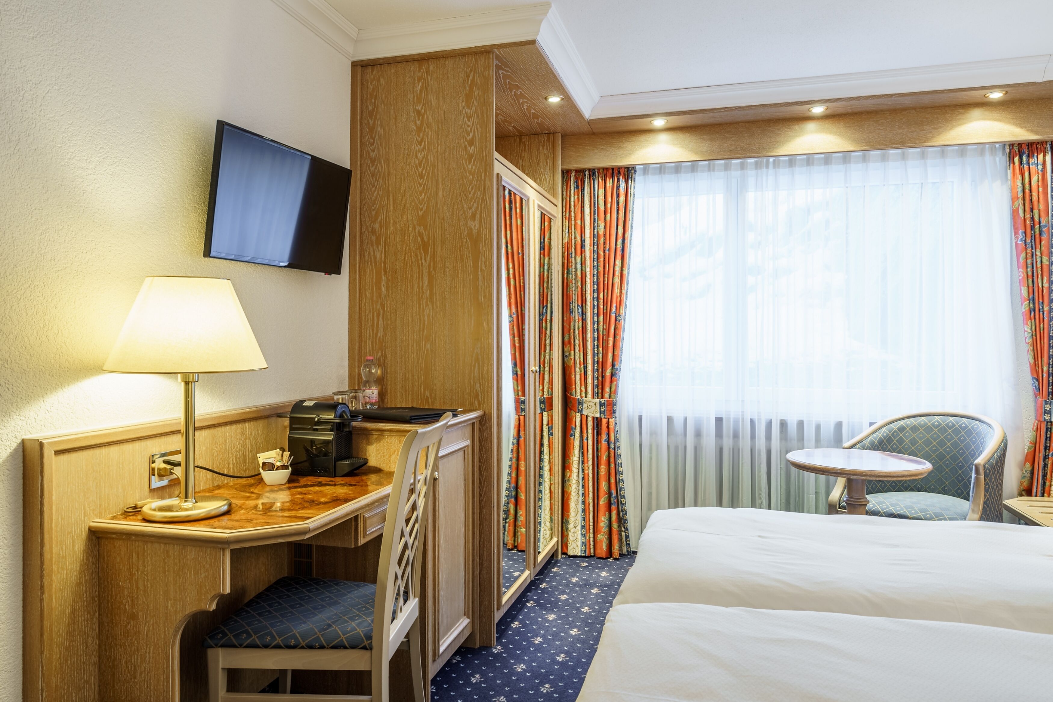 Une chambre d’hôtel avec un lit, un bureau avec une lampe, une chaise, une télévision et une fenêtre avec des rideaux.