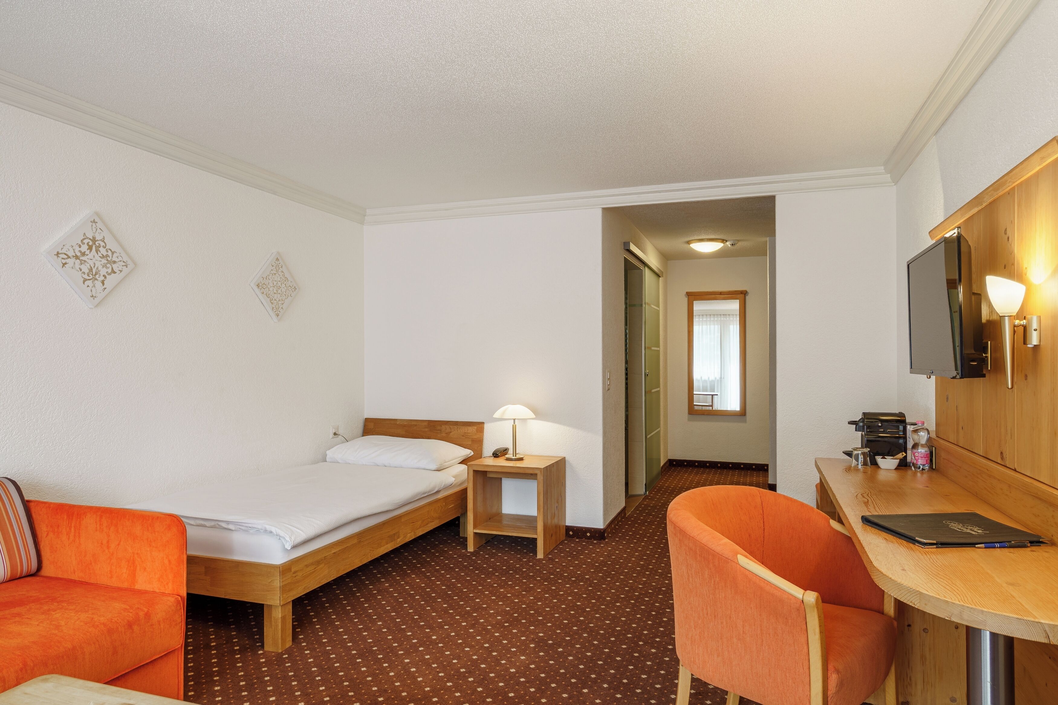 Une chambre d’hôtel avec un lit simple, un bureau avec une chaise, une petite table avec une lampe et un téléviseur.