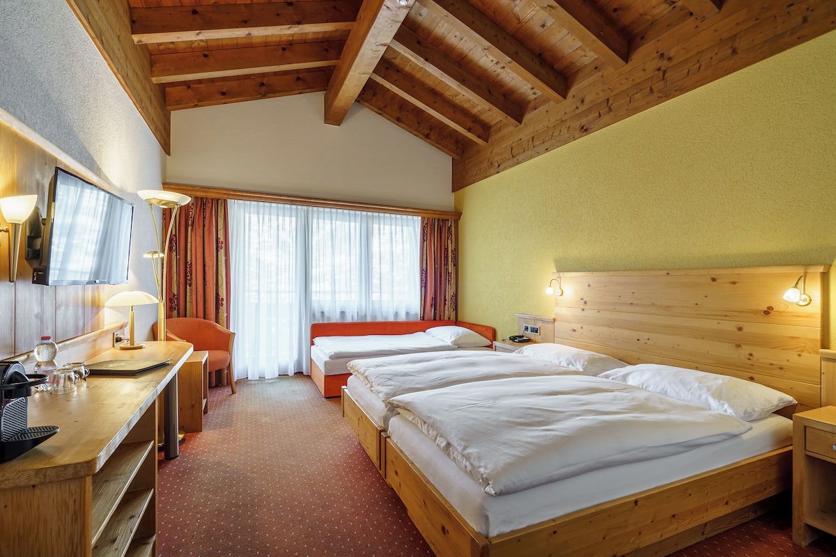 A hotel room with wooden beams, a desk with a lamp, two beds, a chair, and a window with curtains.