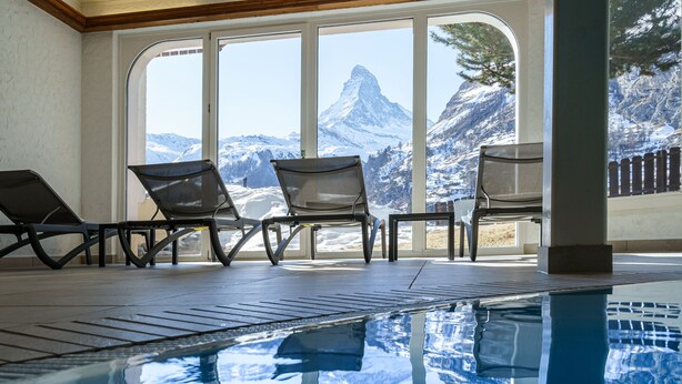 An indoor swimming pool with clear views of a snow-capped mountain and a wooden deck with lounge chairs. - Hotel Alpenroyal (Zermatt)