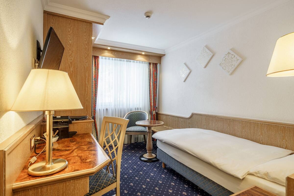 A hotel room with a bed, a desk with a lamp, a chair, a small table, and a window with curtains.