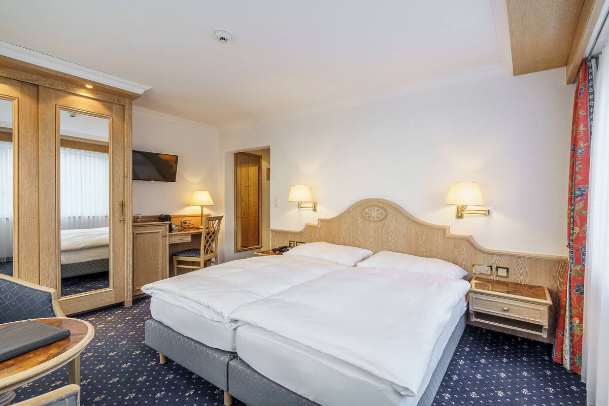 A hotel room with a large bed, two bedside tables with lamps, a desk with a chair, and a wardrobe.