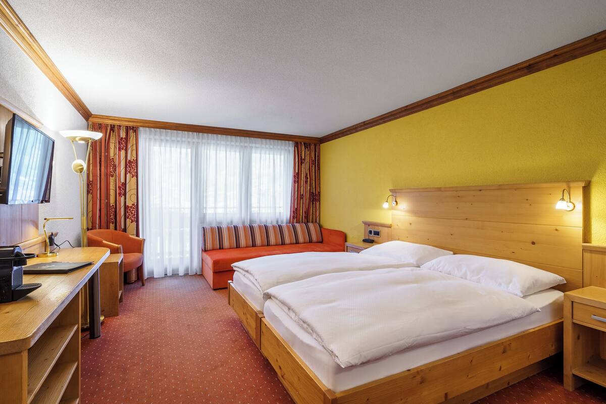 A hotel room with a wooden bed, a desk with a lamp, a sofa, and a television.