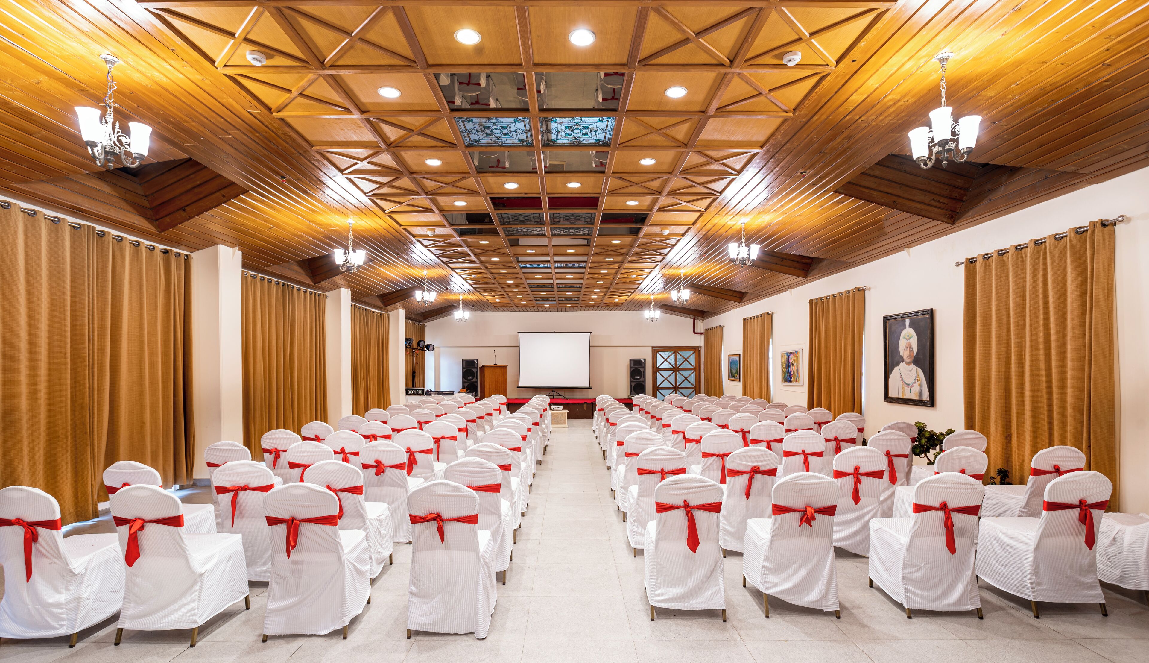 A conference room with rows of white chairs with red sashes, wooden paneling on the ceiling, and a projector screen at the front.