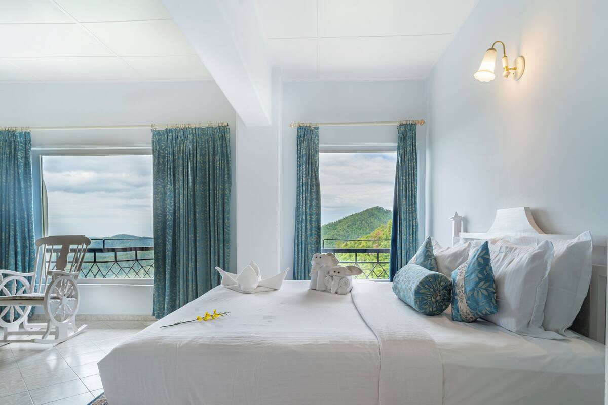 Un dormitorio con una cama grande, dos ventanas con cortinas, una silla y vistas a un paisaje verde.