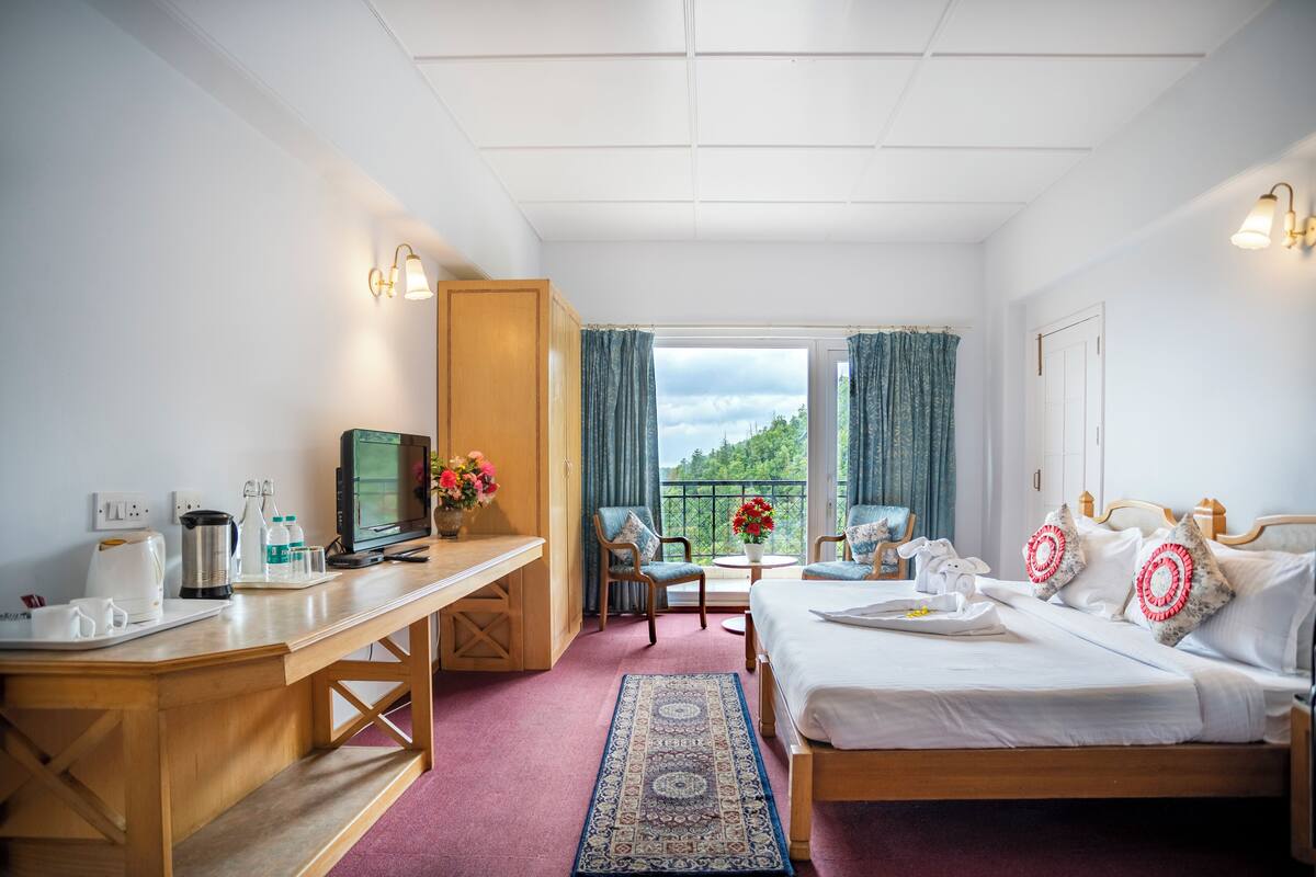 Habitación de hotel con cama, escritorio, silla, televisor y balcón con vistas.