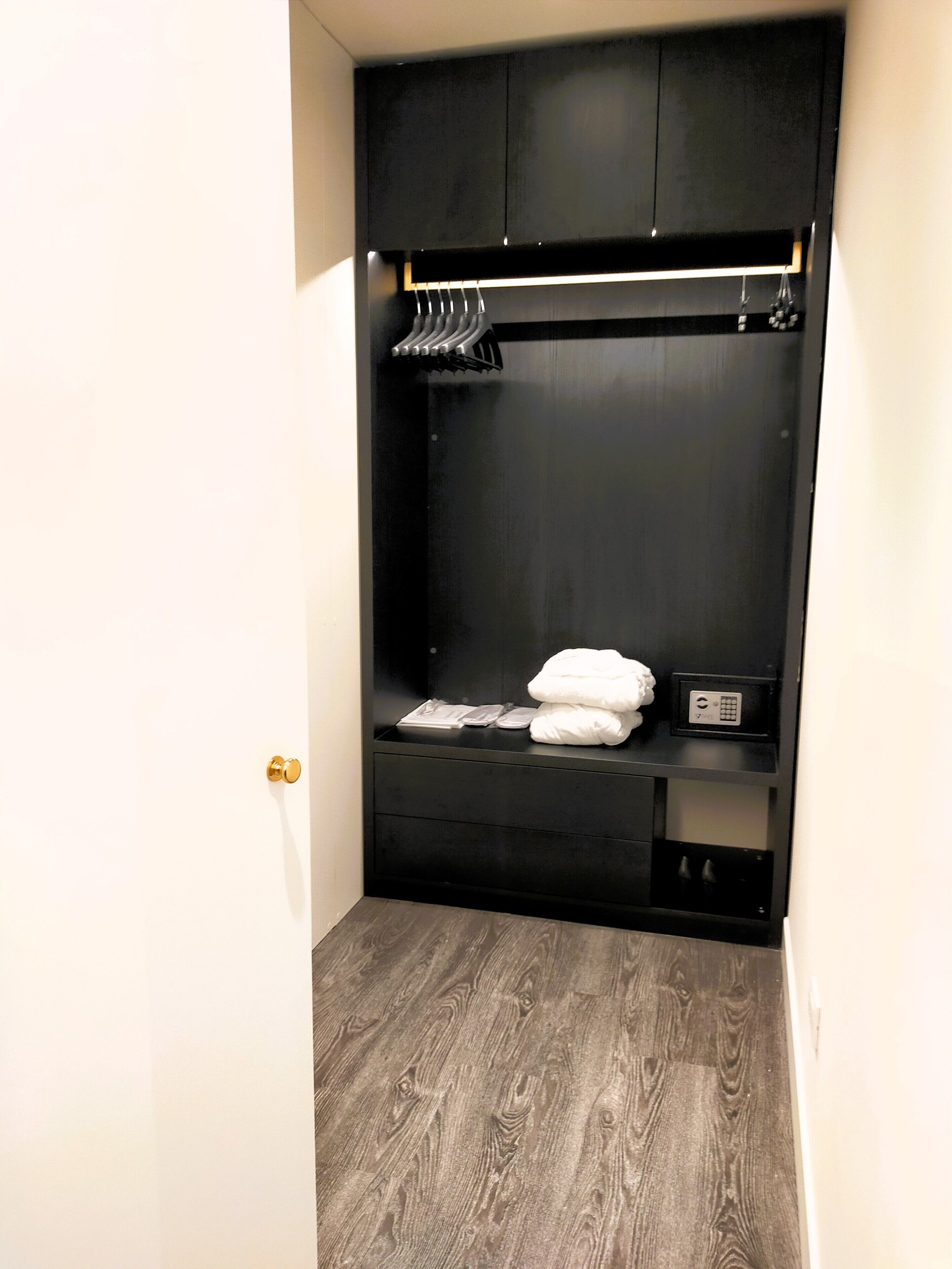 A compact, dark-colored wardrobe with a hanging rack, a shelf, and a small drawer, situated in a room with a wooden floor and white walls.