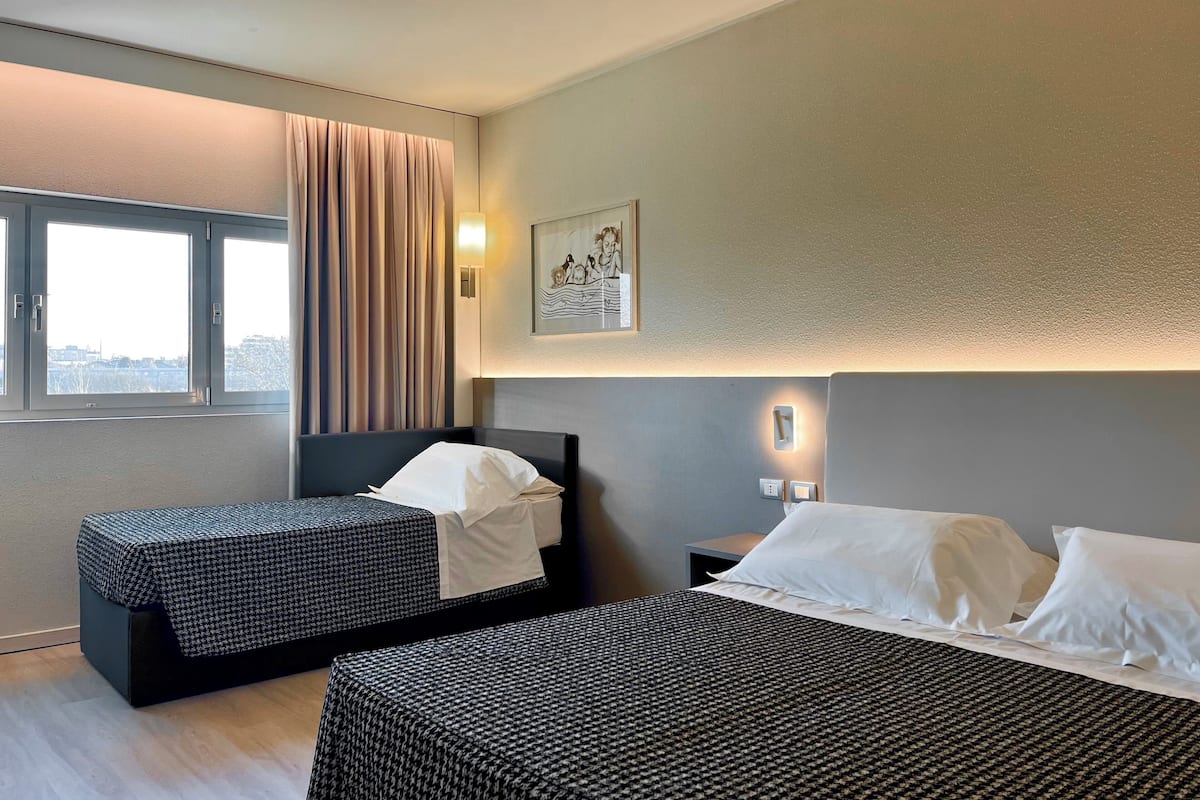 Une chambre d’hôtel avec deux lits, une fenêtre avec des rideaux, un luminaire fixé au mur et un tableau encadré.
