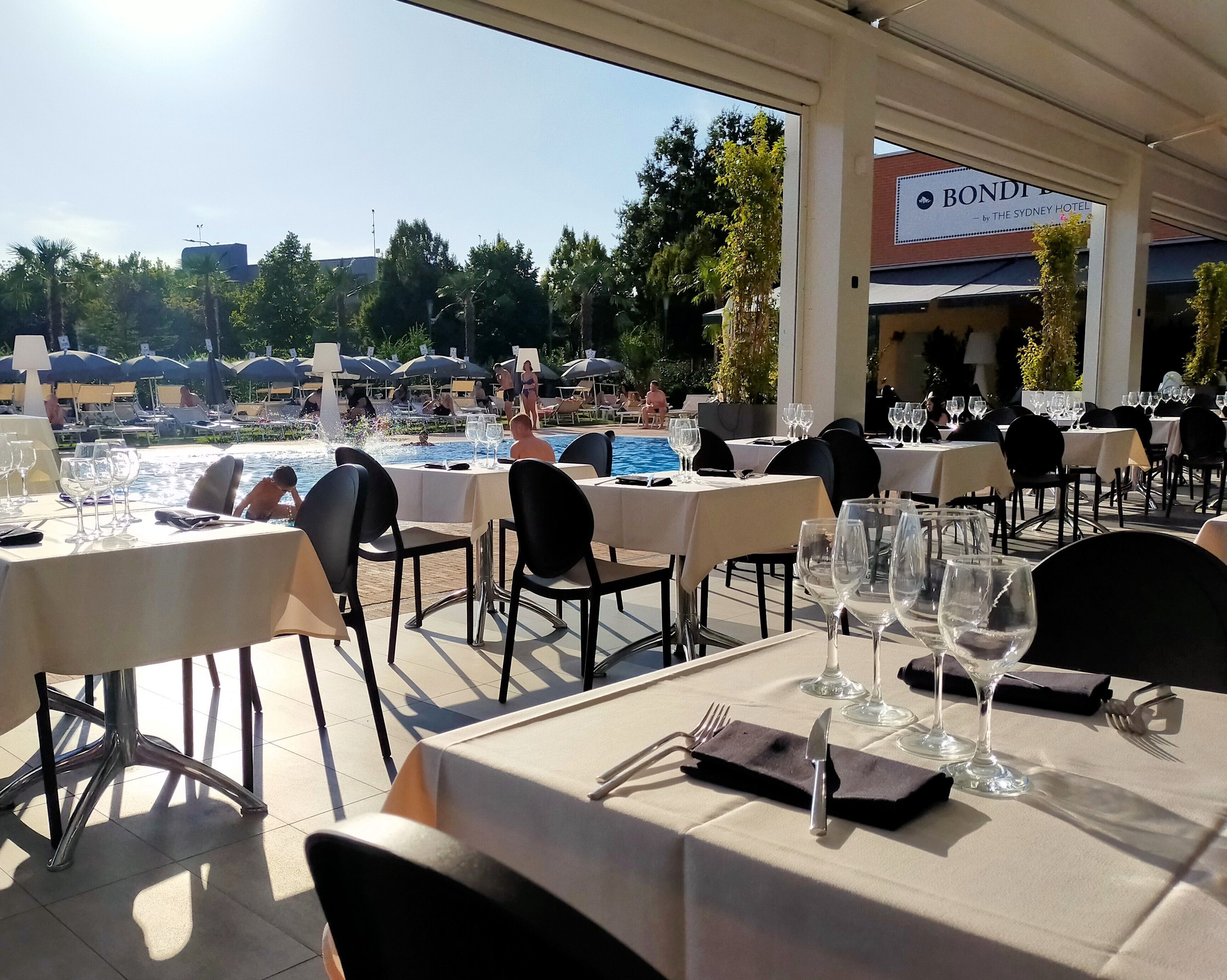 Un área de comedor al aire libre con mesas preparadas para una comida, con vistas a una piscina y un jardín.