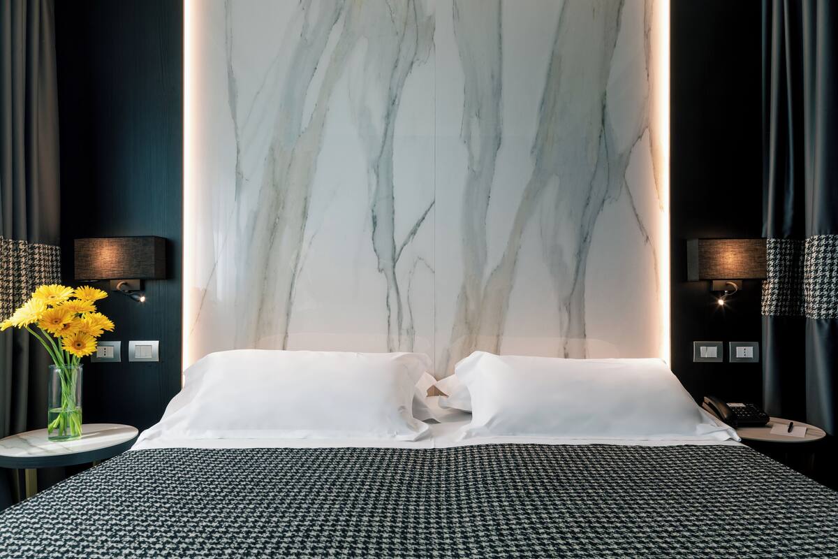 Une chambre d’hôtel avec un grand lit, un mur en marbre, un vase rempli de fleurs jaunes et deux tables de chevet avec des lampes.