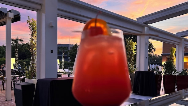 Een glas rode drank met een kers als garnering op een tafel in een terras met uitzicht op een zonsondergang.