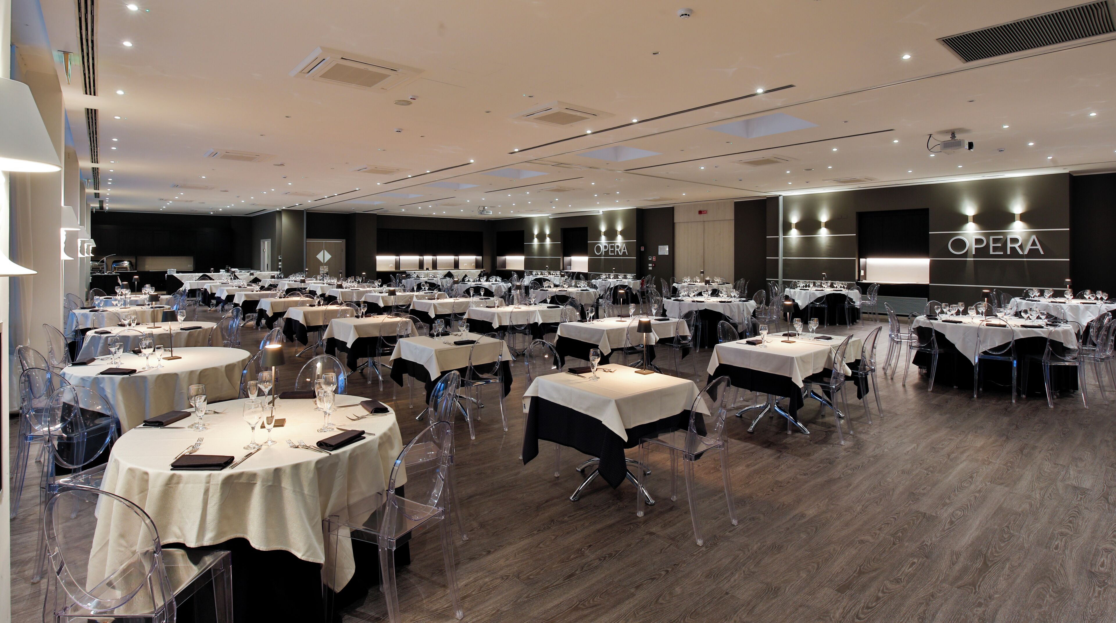 Un'ampia sala da banchetto con numerosi tavoli rotondi apparecchiati per la cena, sedie trasparenti e tovaglie bianche.