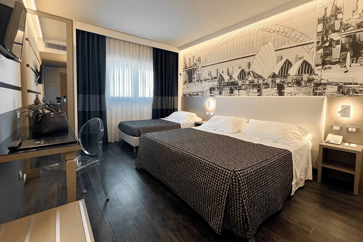 Une chambre d’hôtel avec deux lits, un bureau avec une chaise, un miroir, une fenêtre avec des rideaux et une fresque murale.