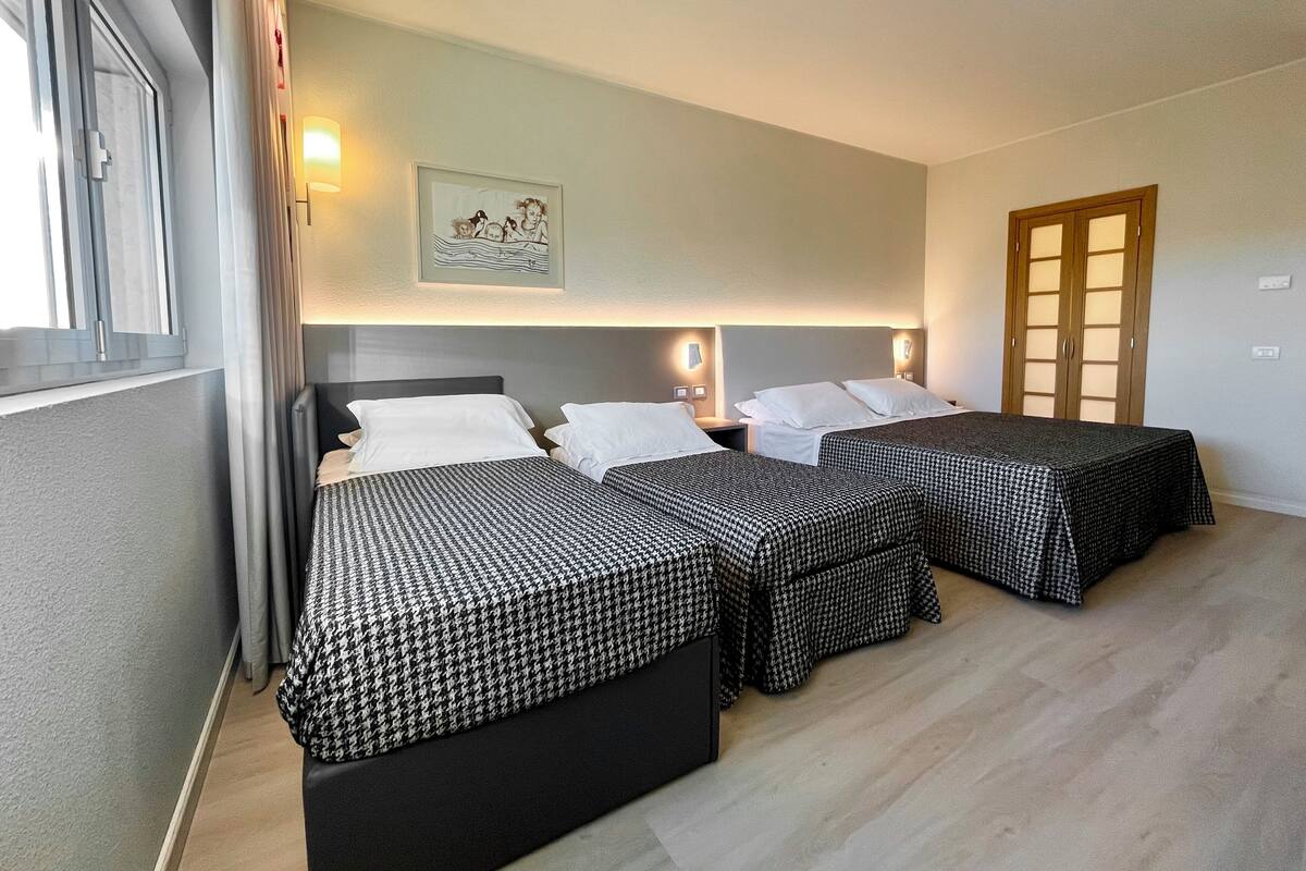 Une chambre d’hôtel avec trois lits, chacun recouvert d’une couverture à carreaux noirs et blancs. Un tableau encadré est accroché au mur au-dessus des lits, et une porte en bois avec des panneaux de verre se trouve à droite.