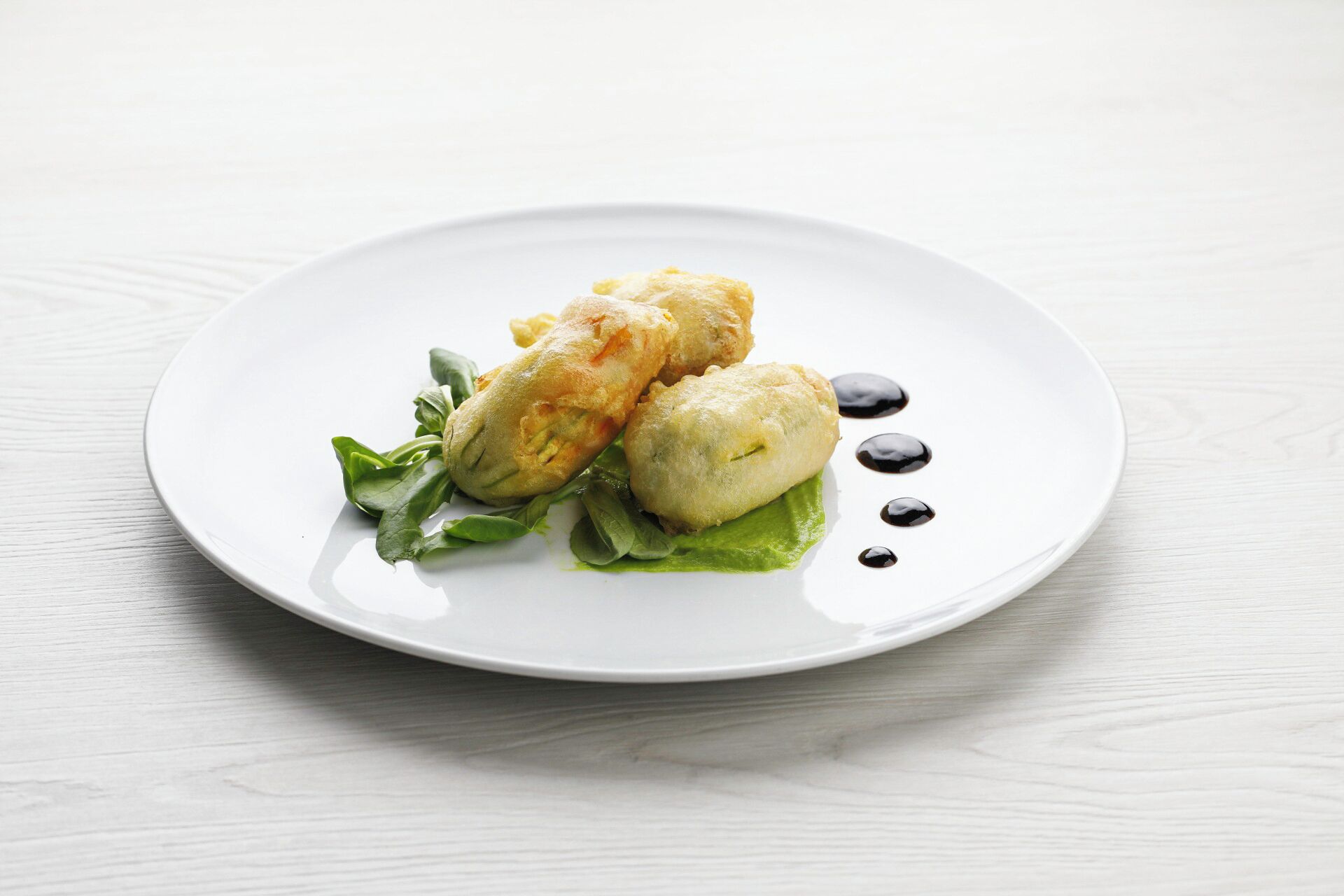 A white plate with two golden-brown pastries on a bed of green leaves, accompanied by a dark sauce.