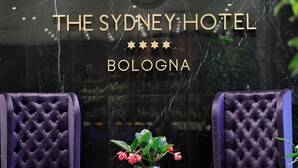 Een lobby met twee paarse, gestoffeerde fauteuils, een bijzettafeltje met een bloevas, en een bordje met de tekst 'The Sydney Hotel Bologna' en een sterrenclassificatie.