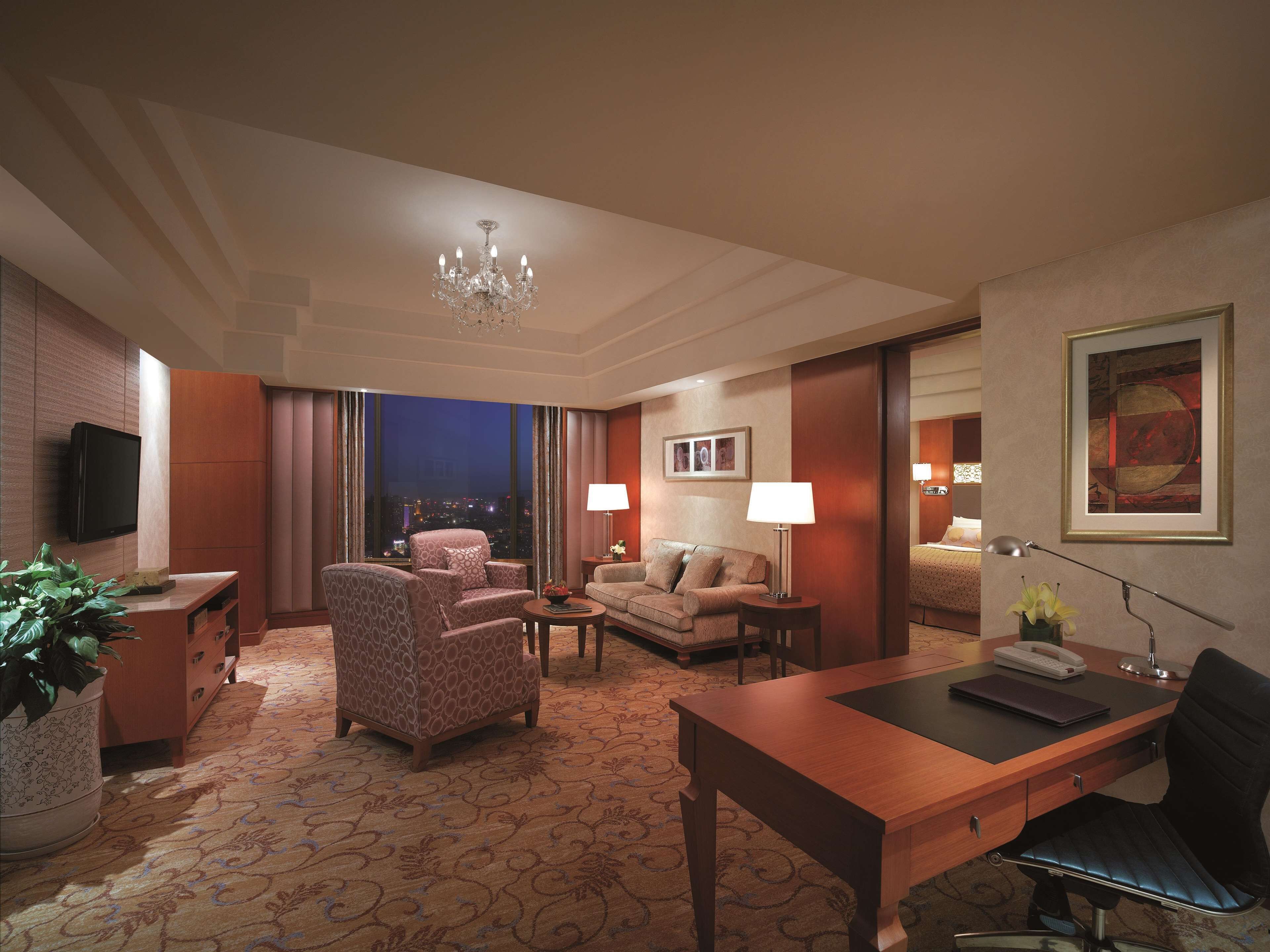 Habitación de hotel con un ventanal grande, un candelabro, un escritorio, un sofá y un televisor.