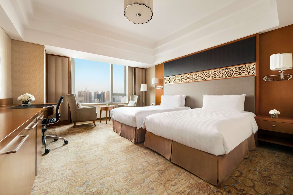 A hotel room with two beds, a desk, a chair, and a view of the cityscape.
