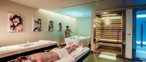 A spa room with two massage tables, a person attending to a client, and artwork on the walls.